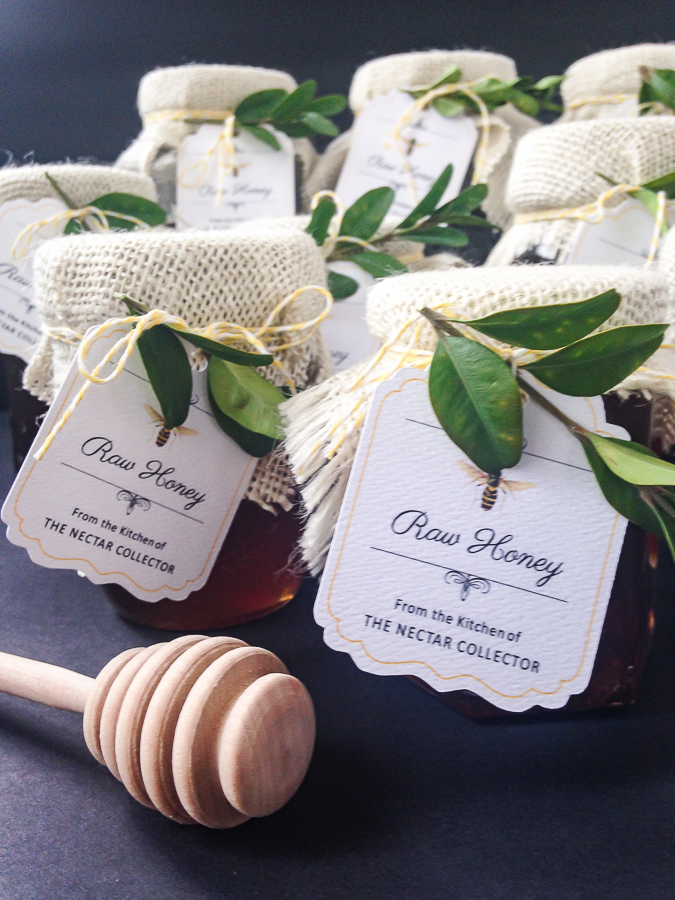 Small glass jars of honey with burlap tops tied with yellow and white striped baker's twine. Tag says "Raw Honey from the kitchen of the Nectar Collector". Sprigs of greenery are tucked in with the twine and a honey stick lays next to the jars.