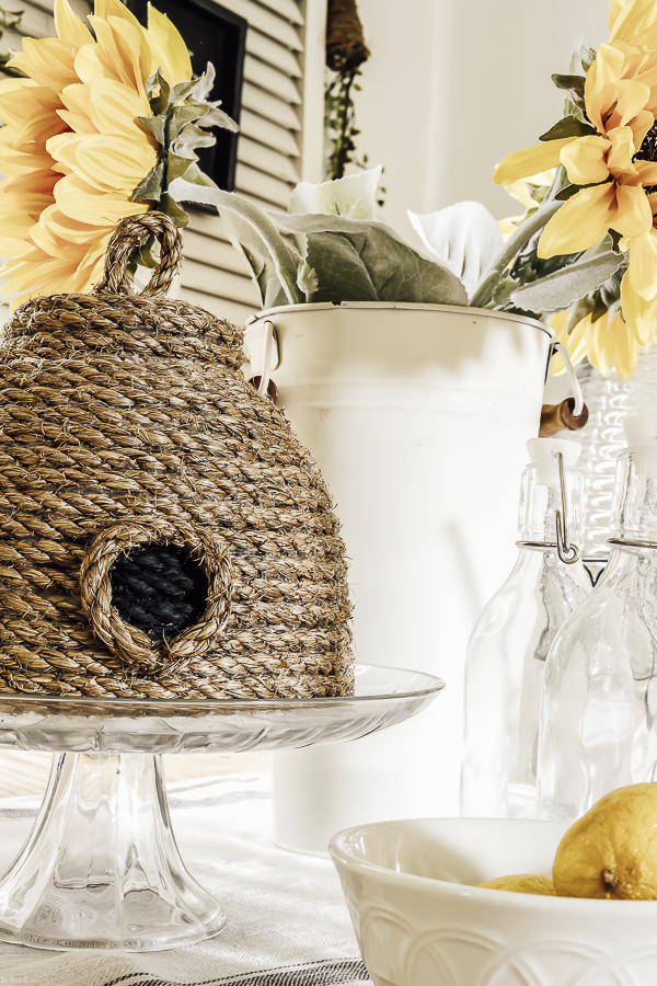 DIY rope bee skep on a glass cake dish in front of a tall tin bucket of sunflowers. A white bowl of lemons and glass jars are in the foregound.