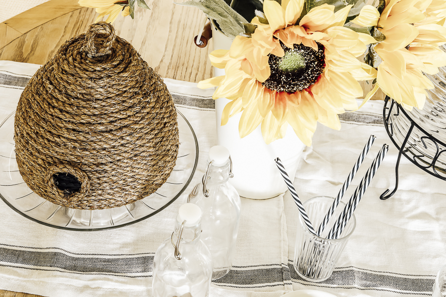 Picture of a DIY bee skep made with twine on a tablescape with yellow sunflowers, glass jars and a glass cup filled with black and white striped straws.