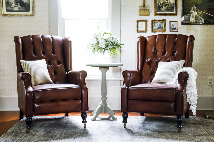 Image of the two made over leather recliners in an English Cottage style living room. They are sitting in front of a window with a light green end table between them that holds a vase of green and white flowers. Behind the chair on the left is a framed vintage portait on the wall papered wall, and behind the chair on the right is a gallery wall of vintage art work in golden frames and an antique paint brush. A floor lamp peaks in from the right side of the frame.