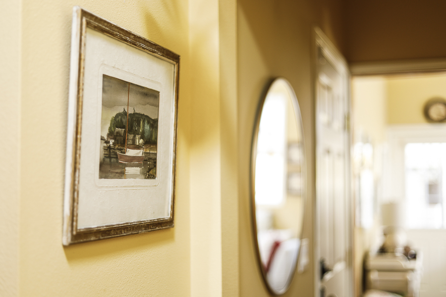 Nautical art of a sailboat is hung in a hallway on a yellow wall. The focus in the rest of the picture toward an exterior doorway is soft and blurred.